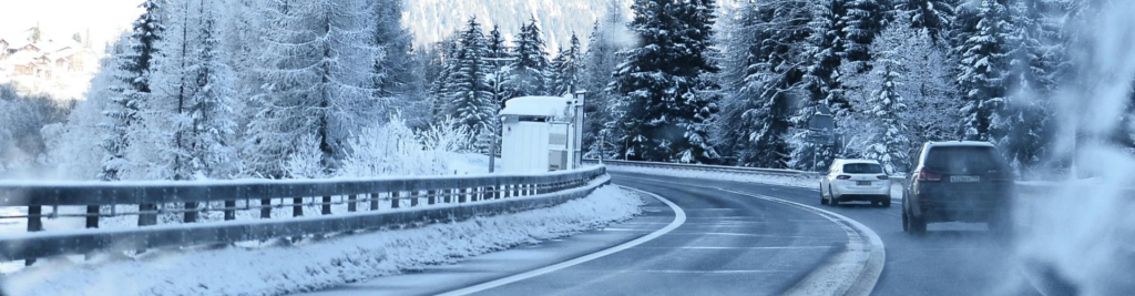 A snowy road lined with evergreen trees, with cars driving around a curve. The sky is overcast and the landscape is covered in snow.